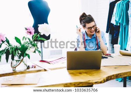 Young successful atelier owner in spectacles having funny mobile conversation in positive atmosphere on smartphone sitting at desktop with sewing equipment and laptop computer in stylish showroom
