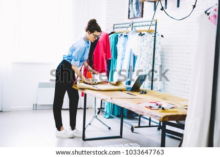 Pencive female entrepreneur owner of fashion shop standing at desktop with design blueprint of stylish clothing.Concentrated talented dressmaker creating new collection of casual apparel in atelier