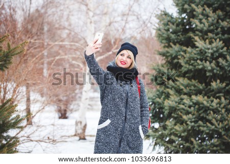A beautiful hipster girl in a gray coat and a black hat makes selfie on the phone outdoors. Portrait of a modern girl who makes selfie, space for text.