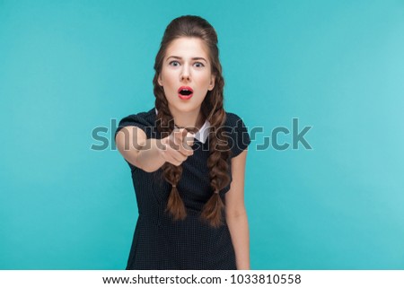 Surprised young woman pointing finger at camera and shocked. Indoor, studio shot on blue background