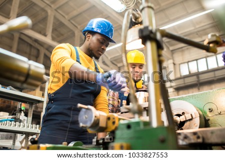 Multi-ethnic team of workers wearing overalls and protective helmets using lathe in order to machine workpiece, interior of spacious production department on background