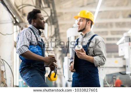 Multi-ethnic team of technicians wearing overalls chatting animatedly with each other while taking coffee break, interior of spacious production department on background