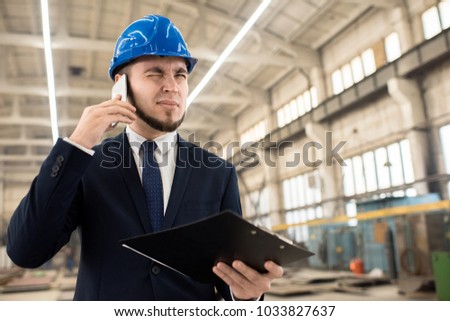 Bearded young entrepreneur wearing hardhat and classical suit talking to his business partner on mobile phone while standing at spacious production department of modern plant, portrait shot