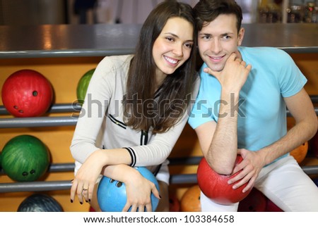 Man and woman sit near shelves with balls and hold balls in bowling club; shallow depth of field