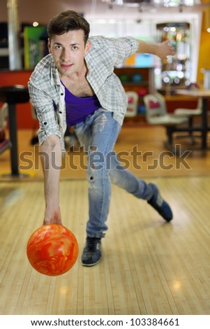 Young smiling man wearing ripped jeans throws ball in bowling; shallow depth of field