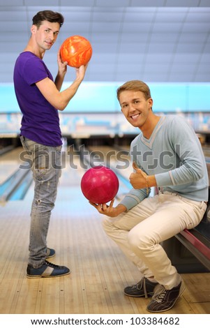 Two young happy men hold balls in bowling club; right man sits and thumb up; left man prepares to throw ball and looks at camera; focus on right man; shallow depth of field