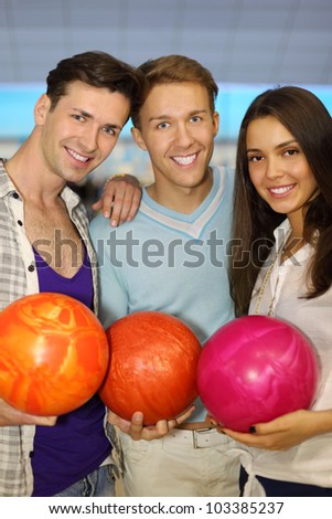 Two young men and beautiful girl hold balls in bowling club; focus on center man; shallow depth of field