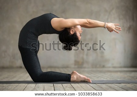 Young woman practicing yoga, doing Preparation for kapotasana, variation of Ustrasana exercise, Camel pose, working out, wearing sportswear, black pants and top, indoor full length, gray wall, studio