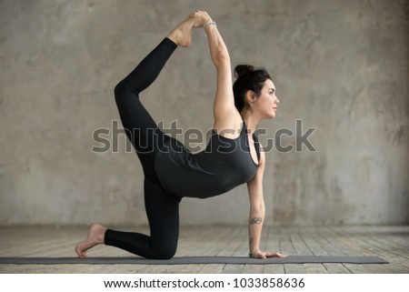 Young woman practicing yoga, doing tiger exercise, Bird dog pose, working out, wearing sportswear, black pants and top, indoor full length, gray wall in yoga studio