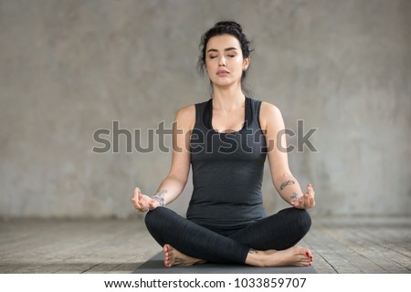 Young sporty woman practicing yoga, doing Sukhasana exercise, Easy Seat pose, working out, wearing sportswear, black pants and top, indoor full length, yoga studio Royalty-Free Stock Photo #1033859707