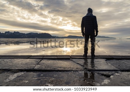 Man standing at the shore, looking at the calm sea. Reflections of the man in the ice on the ground. Mist and fog. Hamresanden, Kristiansand, Norway