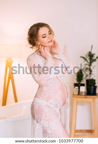 Young, pregnant woman posing in white negligee, background interior in Scandinavian style and bathroom. The photo shows a beautiful belly, the girl smiles cute.
