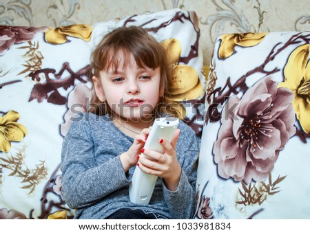 A little girl sits on a couch with a remote and watches TV.