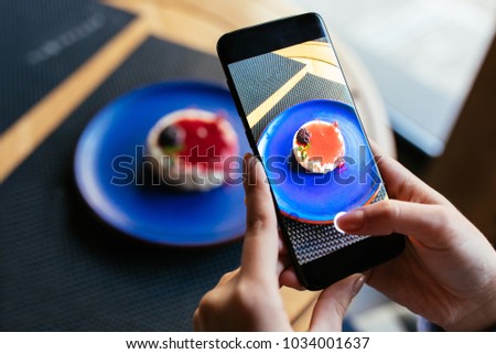Woman using her smartphone to take a photo of cheesecake, while sitting at cafe. Close-up.