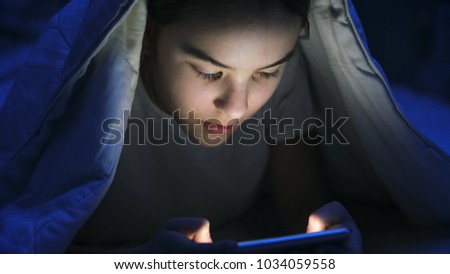 Closeup image of girl in pajamas browsing internet on smartphone under blanket at night