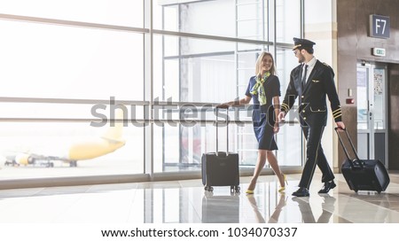 Handsome male pilot and attractive female flight attendant are walking in airport terminal together. Royalty-Free Stock Photo #1034070337