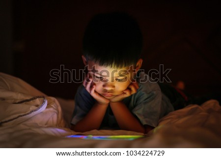 Little asian kid alone watching tablet device, lying on white duvet bed with chin on hands, in background darkness bedroom night time. Colourful bright light from screen reflex on the boy face. Royalty-Free Stock Photo #1034224729