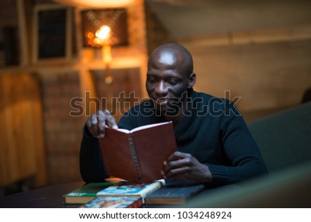 A young African man is reading a book in a room at a table. student, businessman