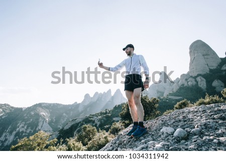 Trendy, fit and strong young man athlete in short running shorts and wind light jacket searches for network or connection on mobile smartphone, or makes photo selfie after workout or hike