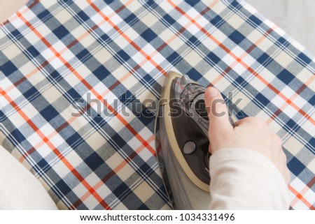 Close up cropped photo woman hand holding iron. Housewife in light clothes ironing checkered shirt, clothing on ironing board with iron. Housekeeping concept. Copy space for advertisement
