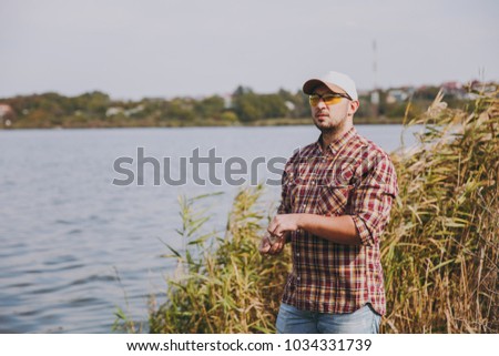 Young unshaven man in checkered shirt, cap and sunglasses looks afar and pulls out of small box maggot bait for fishing against background of lake, shrubs, reeds. Lifestyle, fisherman leisure concept