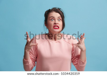 Angry woman looking at camera. Aggressive business woman standing isolated on trendy blue studio background. Female half-length portrait. Human emotions, facial expression concept. Front view.