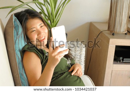 young beautiful and relaxed Asian Chinese woman lying at home living room sofa couch using internet on mobile phone happy and comfortable at modern sweet villa in Summer holidays