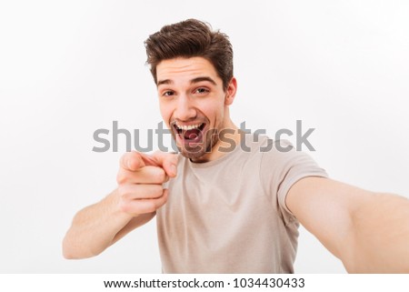 Studio photo of positive man in casual t-shirt and bristle on face smiling and pointing finger on camera while taking selfie isolated over white background