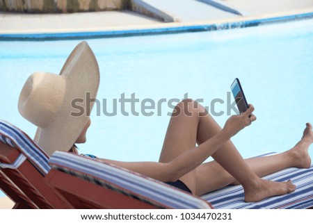Profile view of unrecognizable young woman wearing swimsuit and straw hat lying on deck chair and taking selfie with help of smartphone, blue water surface of outdoor swimming pool on background