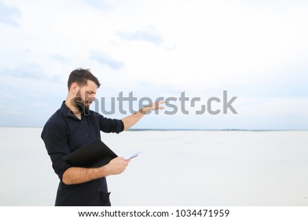 builder man come to deserted place to plan new residential complex, bearded guy standing on sand holding folder with project. Handsome fair-haired concentrated male wearing strict shirt. Concept