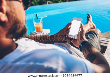 Back view of male blogger holding modern smartphone with mock up blank copy space screen for your advertising information content in hands resting on sun bed with cocktail near blue swimming pool