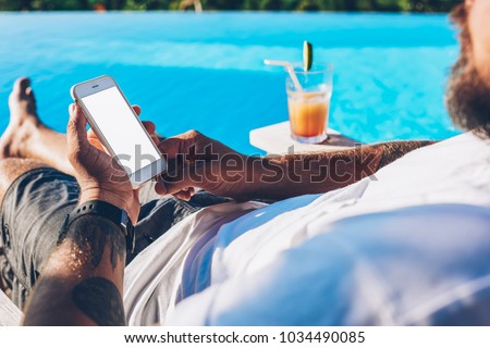 Cropped image of man lying near swimming pool with cocktail and chatting online via high speed internet connected to modern smartphone.Male installing app on mobile phone enjoying summer vacations