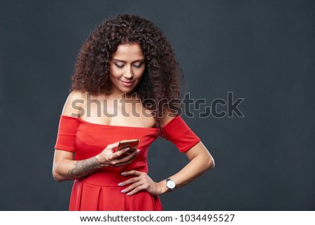 Beautiful curly female in red dress holding smartphone and texting a message