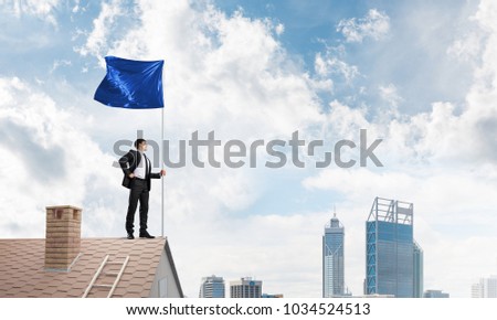 Businessman standing on house roof and holding blue flag. Mixed media