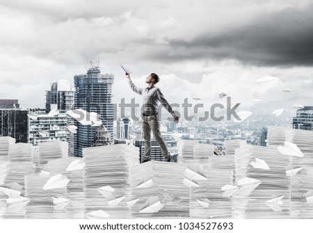 Man in casual wear keeping hand with book up while standing among flying paper planes with cityscape on background. Mixed media.