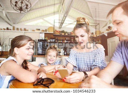  Happy family with two daughters having dinner and using smartphone at restaurant. Family, parenthood, technology concept.

