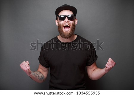 Hipster handsome male model with beard wearing black blank t-shirt with space for your logo or design over gray background Royalty-Free Stock Photo #1034555935