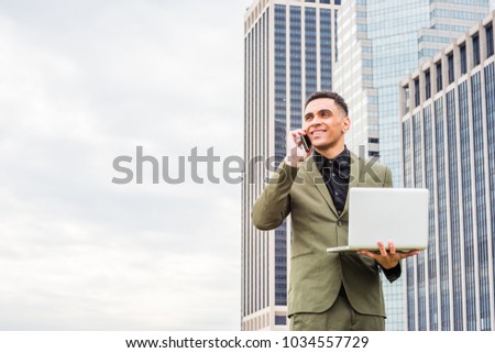 Happy Young American Businessman traveling, working in New York, wearing green suit, standing in front of business district with high buildings, working on laptop computer, calling on cell phone.
