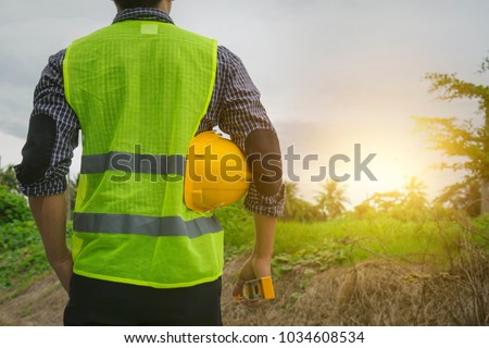 Engineer or architect, wearing green vest, holding yellow safety helmet, water level gauge looking at construction site, empty land with morning sun light Future career, home building construction  Royalty-Free Stock Photo #1034608534