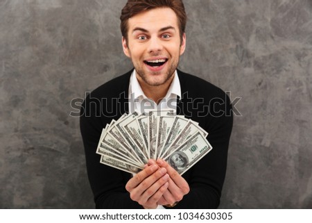 Picture of excited happy young businessman standing over grey wall background holding money looking camera.