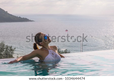 beautiful young woman in white knitted bikini and sunglasses in the rooftop infinity pool against the sea in Phuket