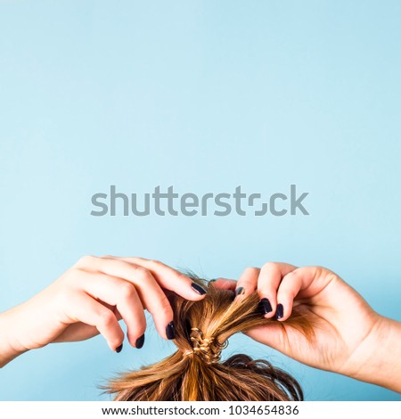 The woman straightens the disheveled bun on her head with her hands with a black manicure. Dark hair is tied with a transparent spiral elastic band. Modern fast hairstyle. Blue background. Copyspace Royalty-Free Stock Photo #1034654836