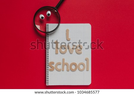 Magnifying glass with eyes looking at "I love school" lettering on a notebook. Red background.