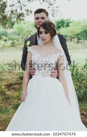 Beautiful wedding couple in the forest. The bride in tulle veil and elegant dress with flowers is hugging the bearded groom in bow tie. Hipster rustic stylish love story outdoors.
