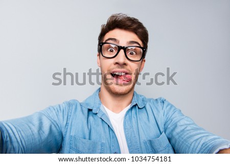 Self portrait of cheerful, positive, handsome guy in casual outfit shooting selfie on front camera of smart phone with two arms, showing tongue out, isolated on grey background