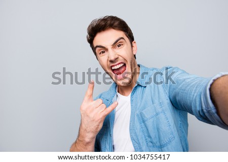 Self portrait of shouting, foolish, individual, attractive, crazy, funny man with wide open mouth, stubble in jeans shirt shooting selfie on front camera, showing rock and roll sign on grey background
