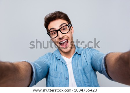 Self portrait of shouting screaming crazy funny man with open mouth, stubble in jeans shirt shooting selfie on front camera with two hands, having video call, isolated on grey background