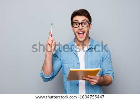 Handsome, attractive, glad, positive, funny guy in glasses with wide open mouth finally found a solution how to make exercise, having raised pen and notebook in hands, isolated on grey background Royalty-Free Stock Photo #1034755447