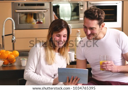 A couple in the kitchen looks at the tablet with the souvenir photos of their holidays or of the past times while having breakfast and smiling happily. Concept of: family, technology, memories.
