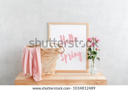 Frame with text HELLO SPRING, woven straw bag and vase with pink rose flowers on a wooden table on a background of light gray walls. Home interior decor. Royalty-Free Stock Photo #1034812489